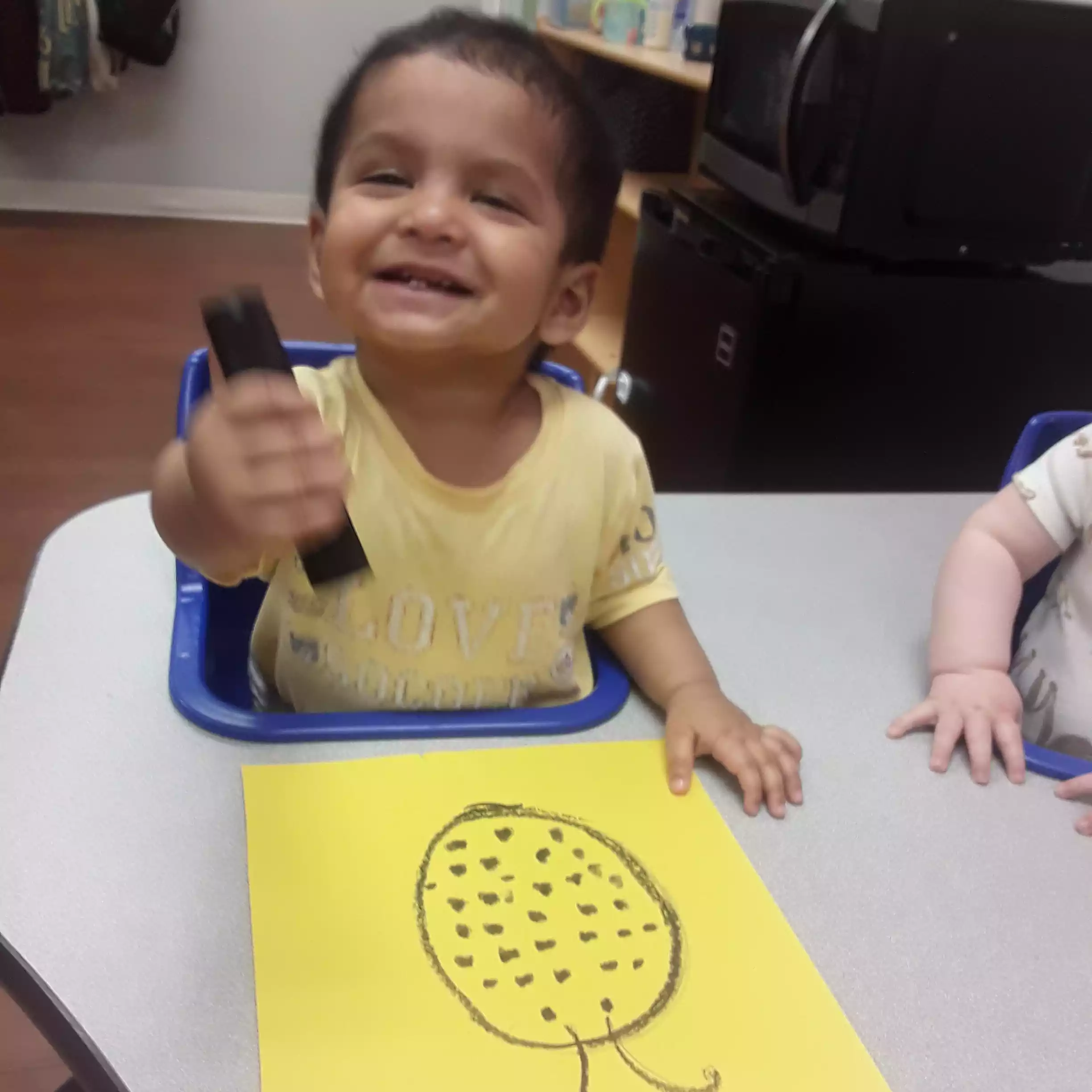Infant Showing Off Ladybug Art Project Happy toddler sitting in an infant program table seat doing a yellow paper craft activity