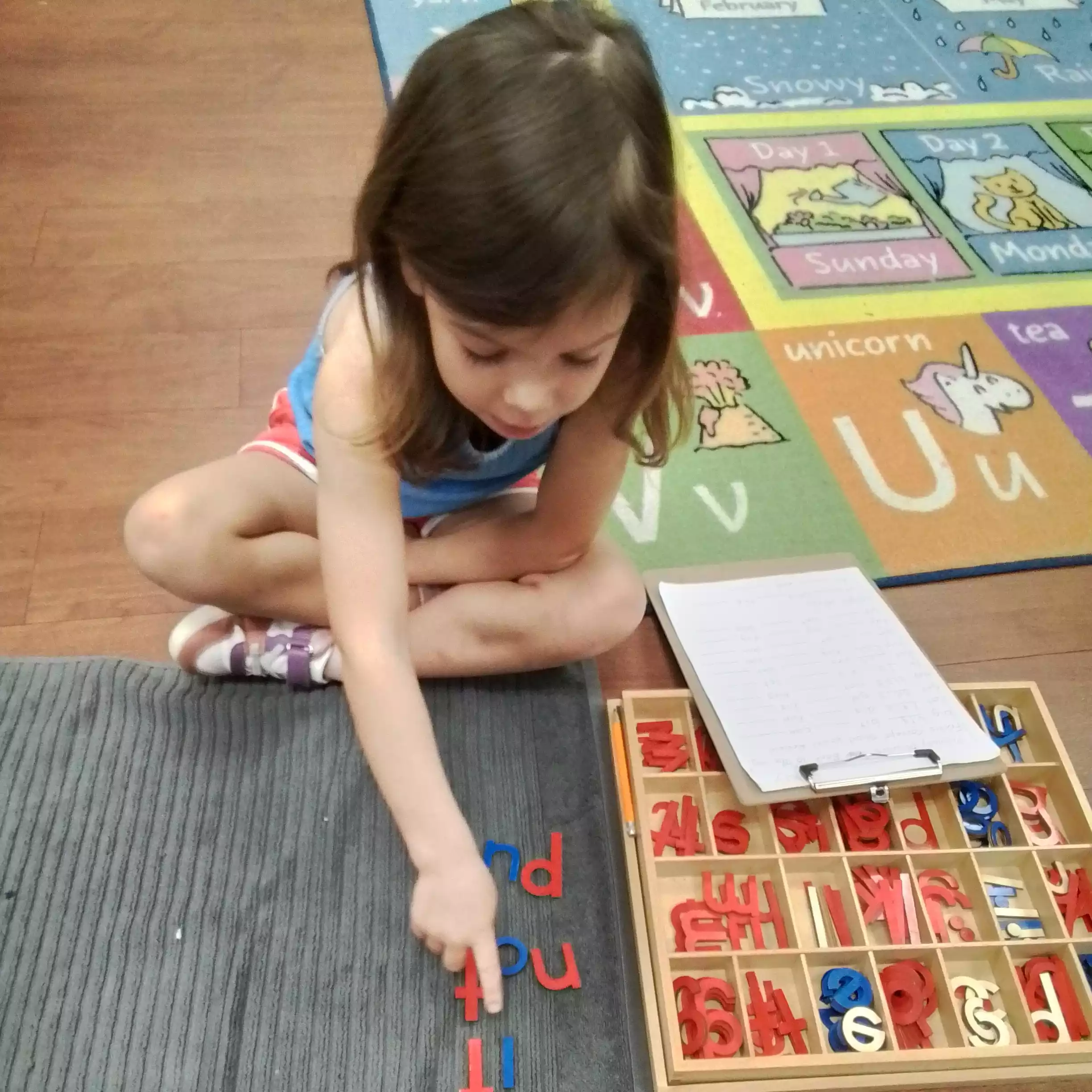 Pinebrook Pre-K Student Practicing Phonics on Reading Mat High-angle view of a young girl pointing at wooden letters spelled out on a mat.