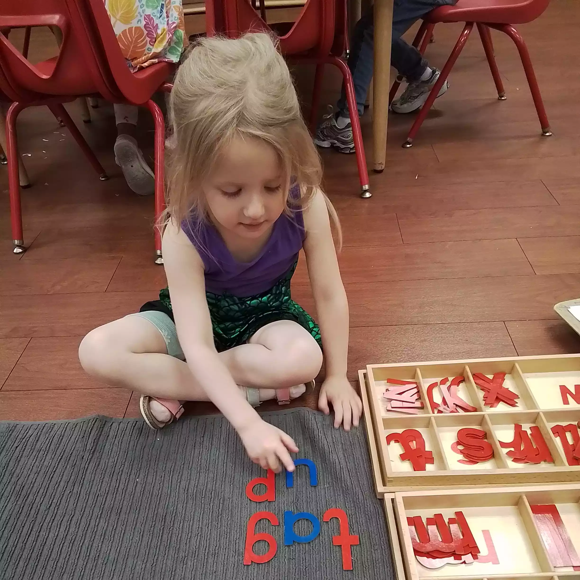 Pinebrook Pre-K Student Practicing Spelling with Cutout Letters Front view of a young girl sitting cross-legged and pointing to spelled words made of wooden letters.
