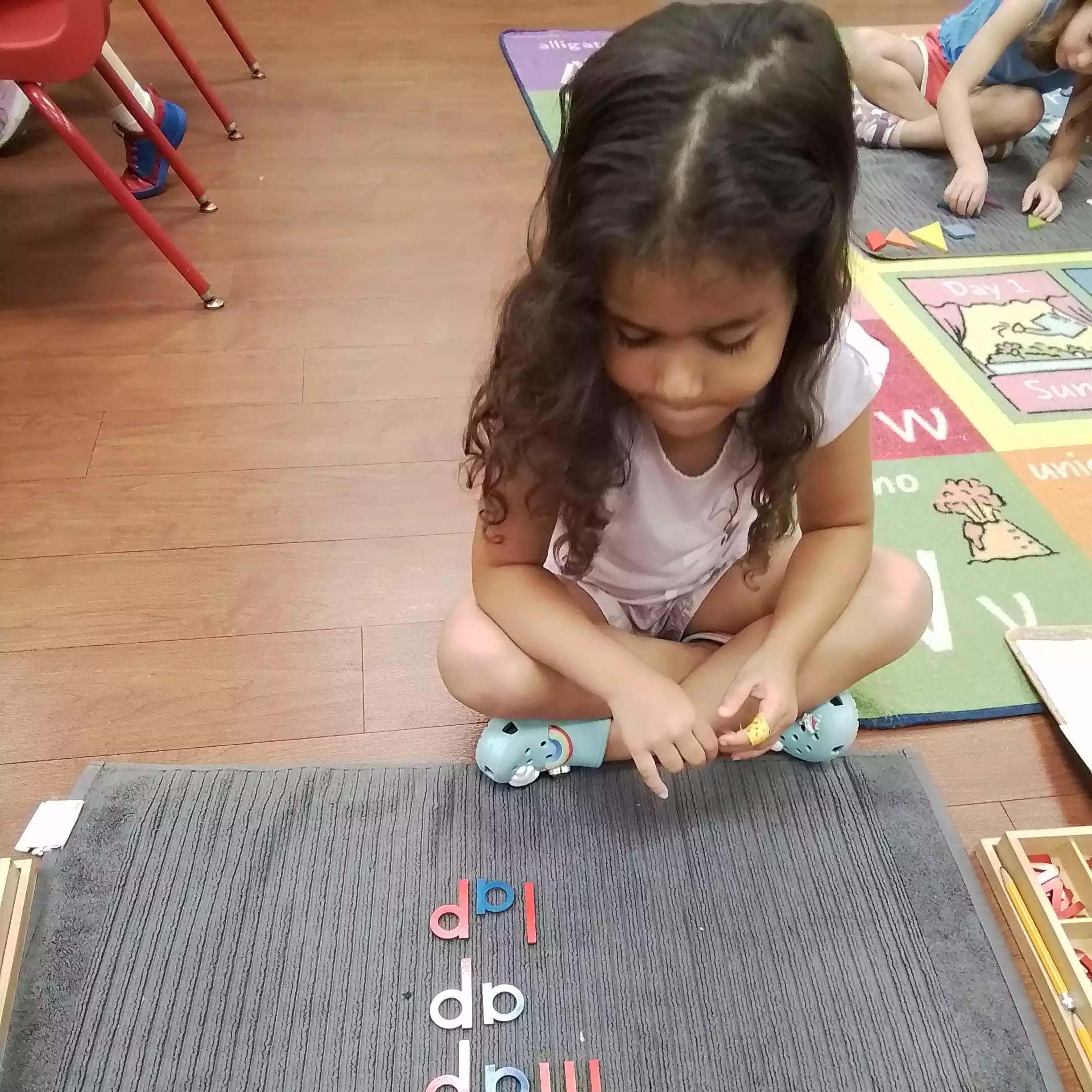 Pinebrook Pre-K Student Studying Wooden Letter Words A young girl sitting cross-legged on the floor looking at wooden letters spelling words on a gray mat.